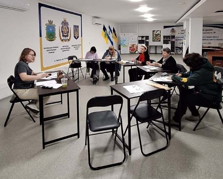 На фото декілька жінок сидять за окремими столами у просторому світлому залі та працюють із паперами. На стіні ліворуч розміщено плакат із гербами, а на задньому плані видно державні прапори України та інформаційні матеріали.