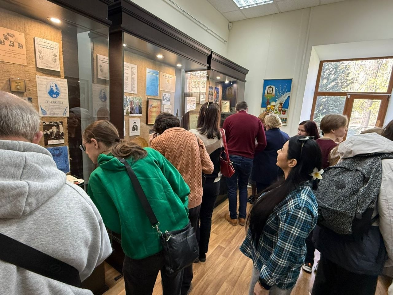 На фото група людей у приміщенні музею чи виставки розглядає експонати у скляних вітринах. У вітринах розміщені фотографії, документи та грамоти. Дехто з відвідувачів нахилився ближче до експозиції, інші стоять поруч. Праворуч видно велике вікно, через яке падає денне світло.
