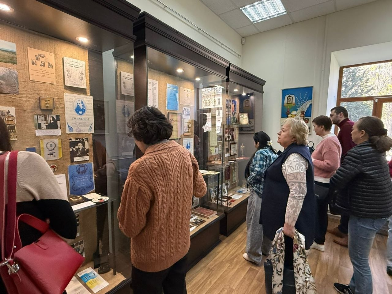 Відвідувачі музею розглядають експозицію, розміщену у високих темних вітринах зі склом. Усередині вітрин представлені архівні документи, старі світлини та грамоти. Люди в повсякденному одязі — светрах та жилетах — уважно вивчають матеріали. Зала світла, з ламінатом на підлозі та великим вікном праворуч, за яким видно дерева.