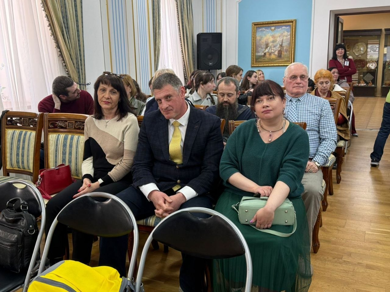 На фото група людей сидить у залі на урочистому заході. На передньому плані — жінка у бежевому джемпері, чоловік у класичному костюмі з жовтою краваткою та жінка в зеленій сукні. Позаду видно інших гостей, інтер'єр з картиною на блакитній стіні та декоративні штори на вікнах.