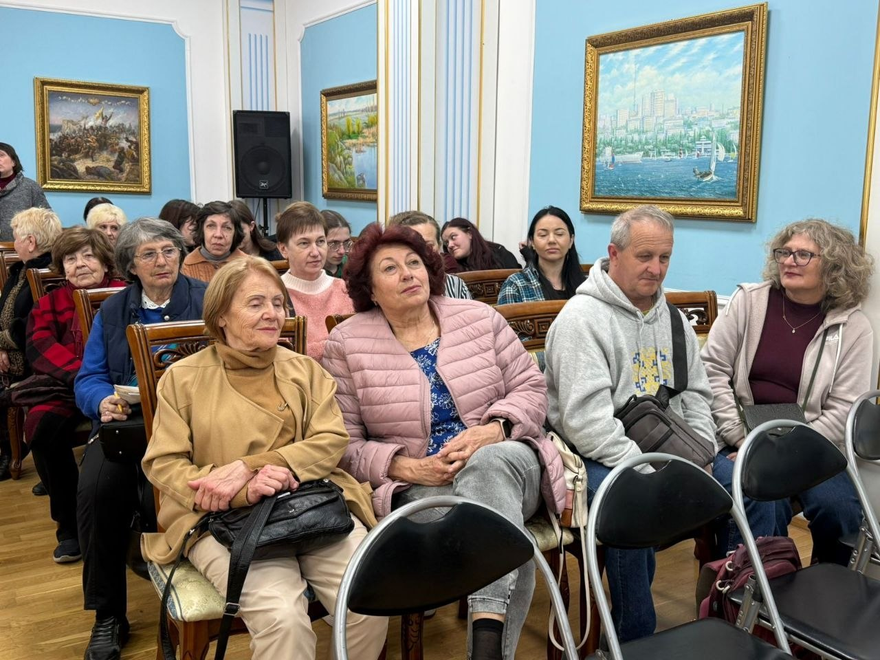 Група людей сидить у залі під час заходу. На передньому плані — жінки у світлому верхньому одязі та чоловік у сірому худі. Блакитні стіни приміщення прикрашені картинами в золотистих рамах, на задньому плані видно інших слухачів та музичну колонку.