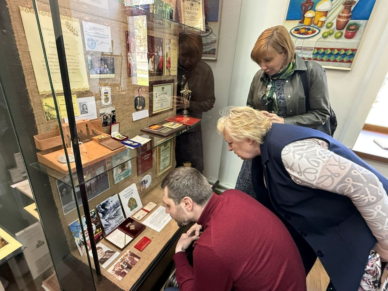 На фото група людей у музеї уважно розглядає експонати у скляній вітрині. Усередині представлені документи, світлини, нагороди та книжки. Одна жінка та чоловік у бордовому светрі нахилилися до нижніх полиць, ще одна жінка стоїть поруч. На стіні праворуч висить картина з натюрмортом.