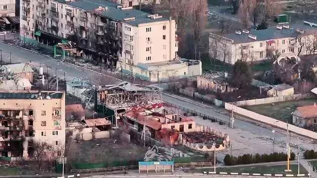 Панорамний вигляд житлового кварталу зі значними руйнуваннями. Багатоповерхові будинки мають сліди пожеж і вибиті вікна, а одноповерхові споруди поряд майже вщент зруйновані.