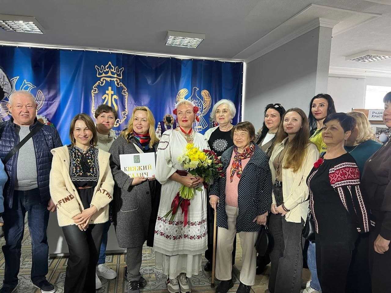 Група людей у приміщенні на фоні великого синього банера з гербом. У центрі стоїть жінка в традиційній вишитій сукні з букетом квітів, а поруч — жінка з табличкою «Княжий Галич».