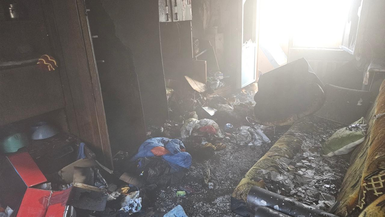 На фото зображено наслідки пожежі в житловому приміщенні. Кімната вкрита шаром попелу та кіптяви, меблі обвуглені, а на підлозі розкидані сильно пошкоджені речі.