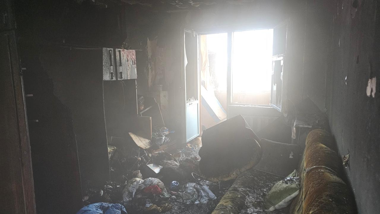 На фото зафіксовано наслідки сильної пожежі в квартирі. Кімната повністю вигоріла: стіни та меблі вкриті чорною кіптявою, на підлозі лежать купи згорілих речей і сміття. Крізь розбите вікно та балконні двері в задимлене приміщення пробивається яскраве денне світло.