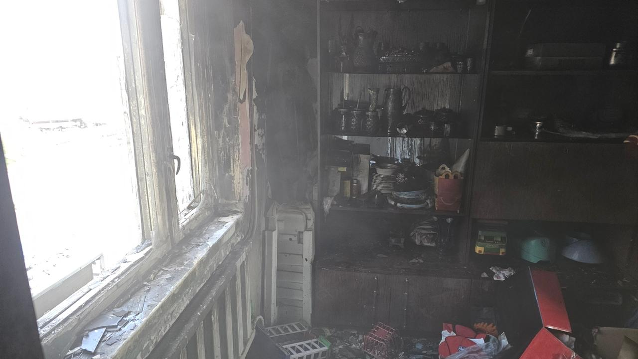 Вигляд зсередини згорілого приміщення. Стіни та меблі повністю вкриті чорною кіптявою, на полицях стоїть обвуглений посуд та побутові речі. Ліворуч видно пошкоджене вікно з обгорілою рамою, крізь яке проходить світло.