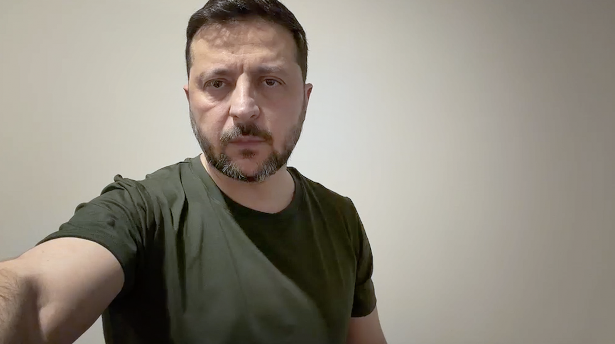 На фото чоловік із темним волоссям та бородою у футболці кольору хакі стоїть на фоні однотонної світлої стіни. Він дивиться прямо в кадр із серйозним виразом обличчя.