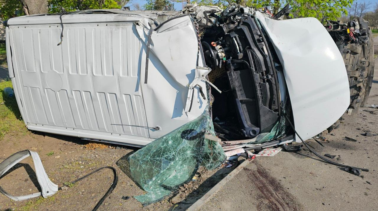 Білий мікроавтобус лежить на боці після серйозної аварії. Передня частина автомобіля повністю розтрощена, лобове скло розбите, а деталі кузова сильно деформовані.