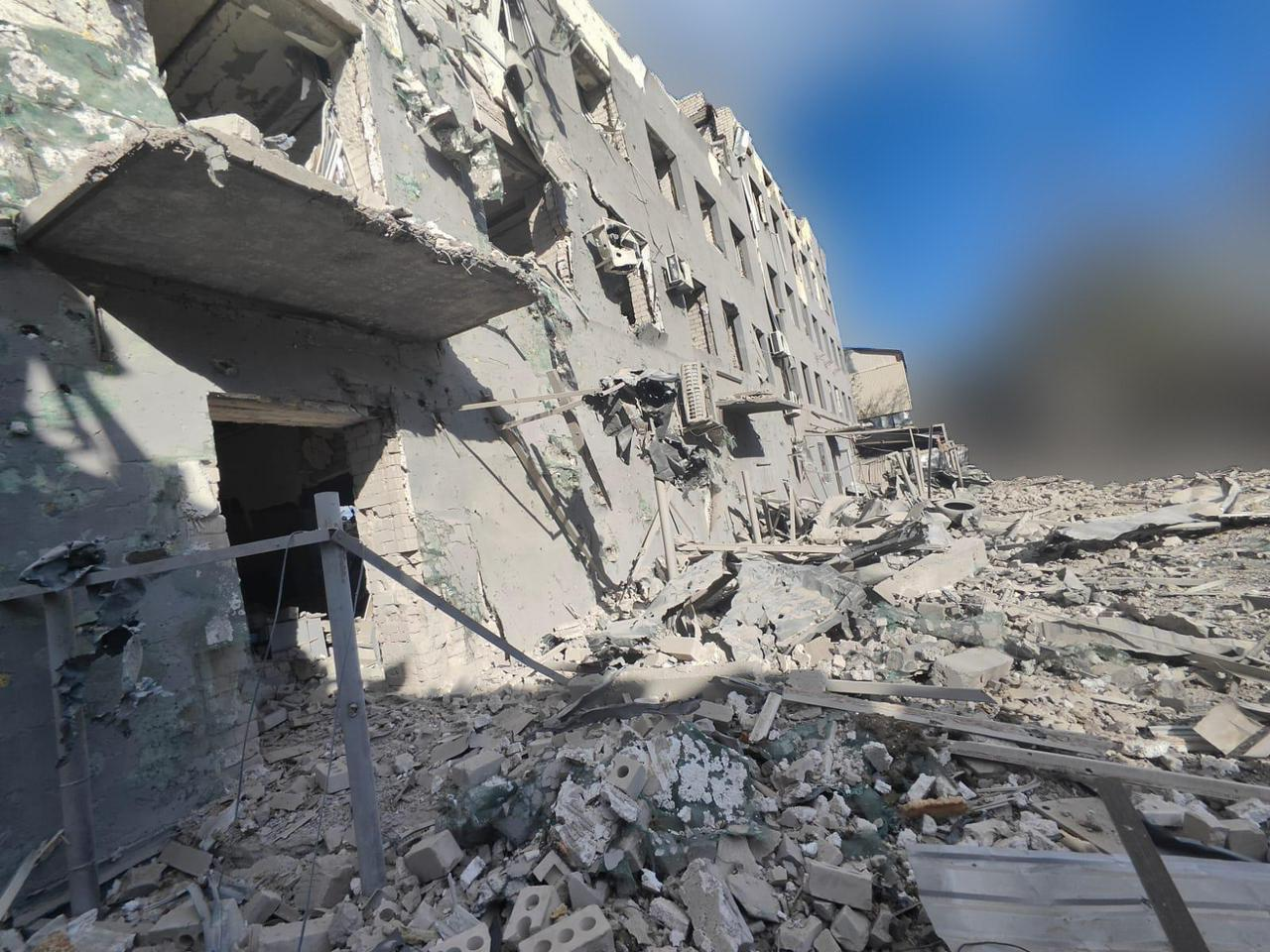 Багатоповерхова будівля зі значними руйнуваннями внаслідок обстрілу. Фасад посічений уламками, вікна вибиті, а перед будинком лежать купи будівельного сміття та бетону.