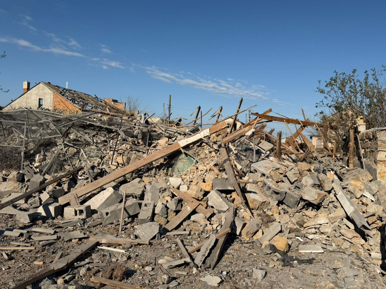 На передньому плані — велика купа будівельного сміття, що складається з розбитих бетонних блоків, цегли та дерев'яних балок. На задньому плані видно напівзруйновану будівлю з пошкодженим дахом під ясним синім небом.