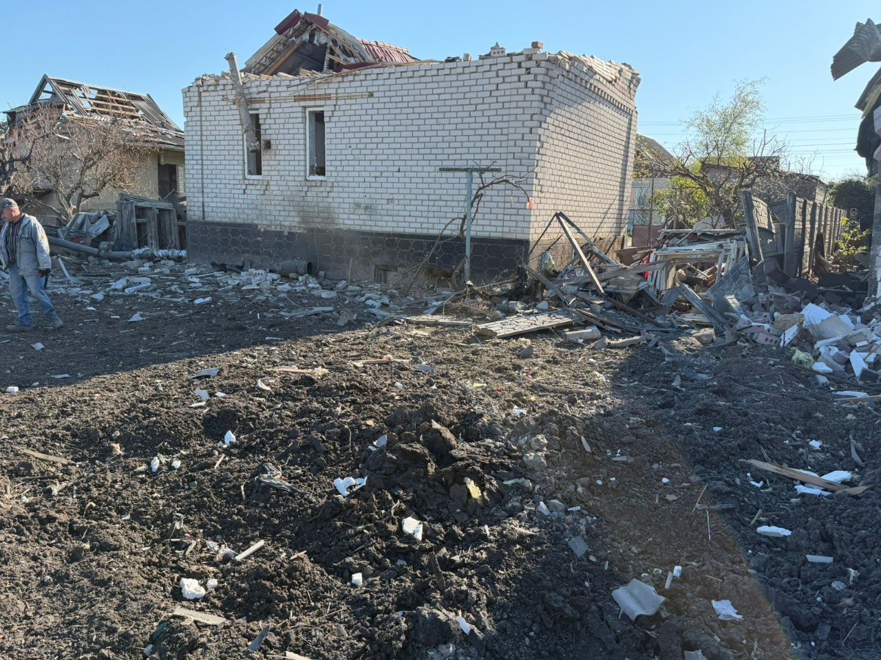 На подвір'ї приватного сектору видно наслідки руйнувань: цегляний будинок з частково обваленою покрівлею та стінами, а поруч — вирва у землі та розкидані уламки будівельних конструкцій.