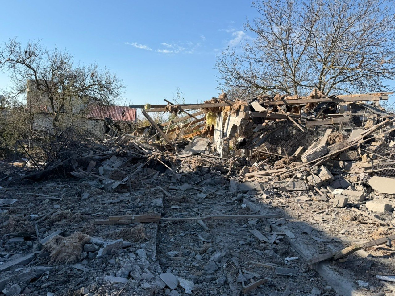 На знімку зафіксовано вщент зруйновану будівлю, від якої залишилася лише купа будівельного сміття, потрощених дерев'яних балок та уламків стін. Навколо місця руйнування стоять голі дерева на тлі ясного неба.