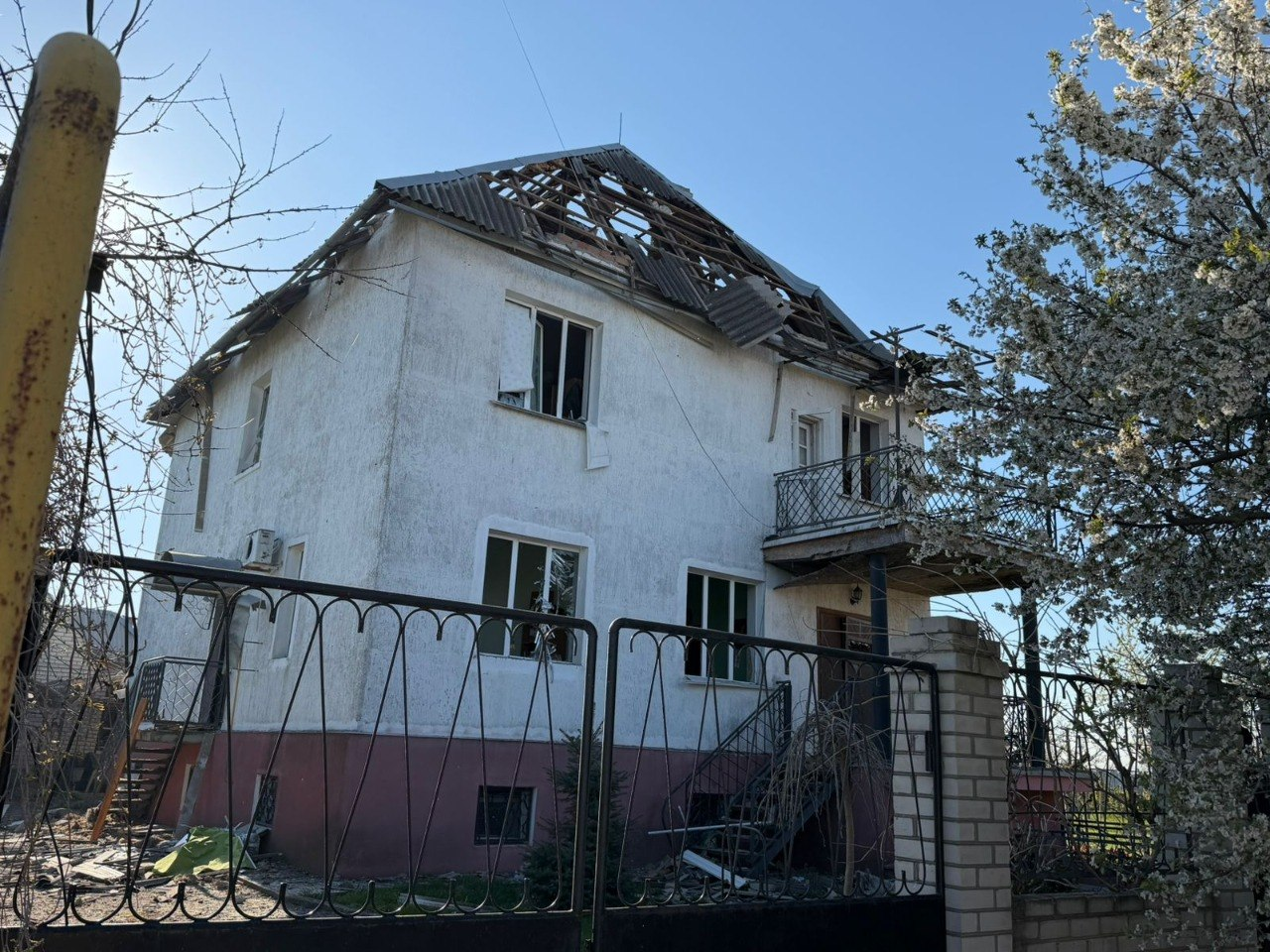 Двоповерховий житловий будинок з пошкодженим дахом, де видно дірки в шиферному покритті. У будівлі вибиті вікна, а на передньому плані — чорний металевий паркан та квітуче дерево поруч із входом.
