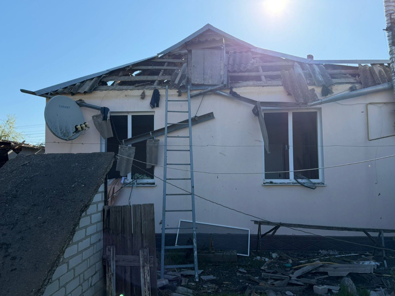 Одноповерховий житловий будинок зі значними пошкодженнями даху та вибитими шибками. До понівеченої покрівлі приставлена металева драбина, а навколо будівлі розкидані уламки шиферу та будівельне сміття.
