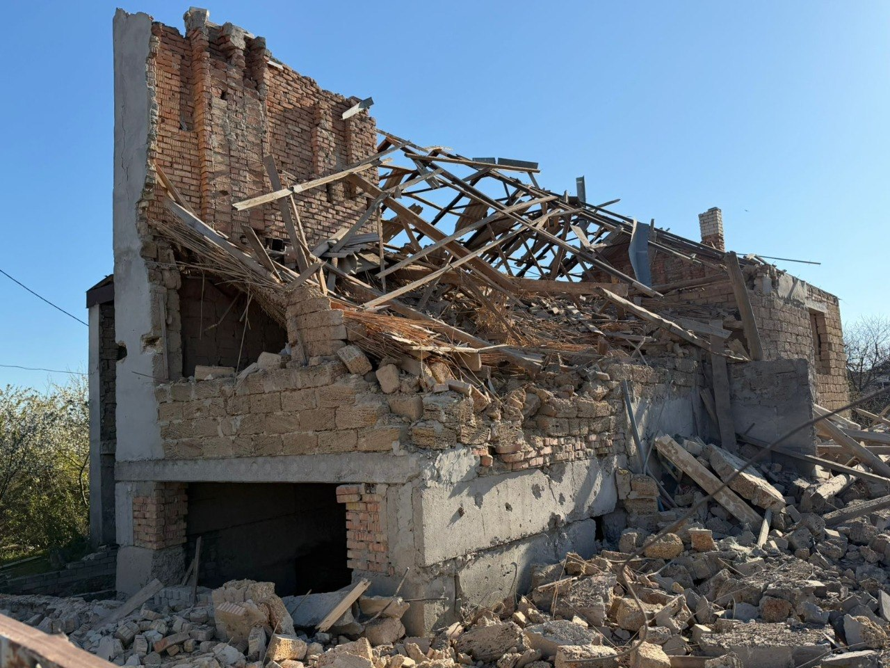 Залишки зруйнованої двоповерхової цегляної будівлі з повністю обваленим дахом та стінами. Повсюди лежать купи каміння, балок та розтрощеної цегли на фоні безхмарного неба.