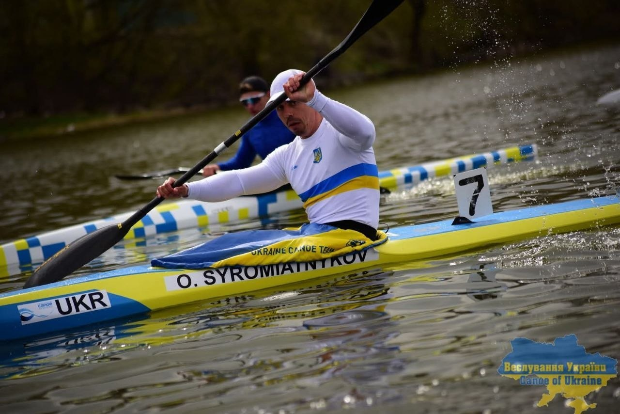 Двоє веслувальників на байдарках під час змагань або тренування на річці. Спортсмен на передньому плані у білій формі з українською символікою активно працює веслом, перебуваючи в жовто-блакитному човні з написом «UKR».