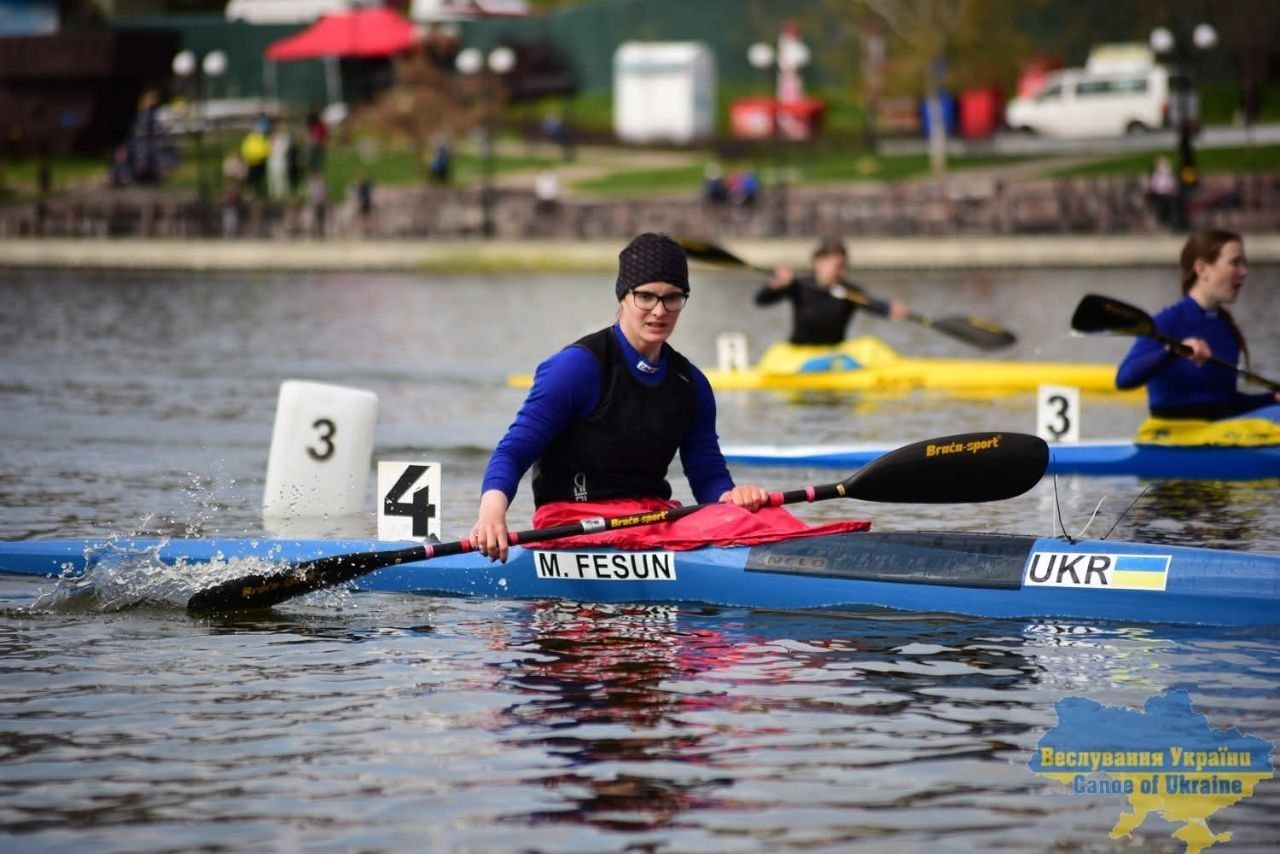 Кілька веслувальниць на байдарках під час змагань на відкритій воді. Спортсменка на передньому плані у синьому одязі та чорній шапці сидить у блакитному човні з написом «UKR» та тримає весло.