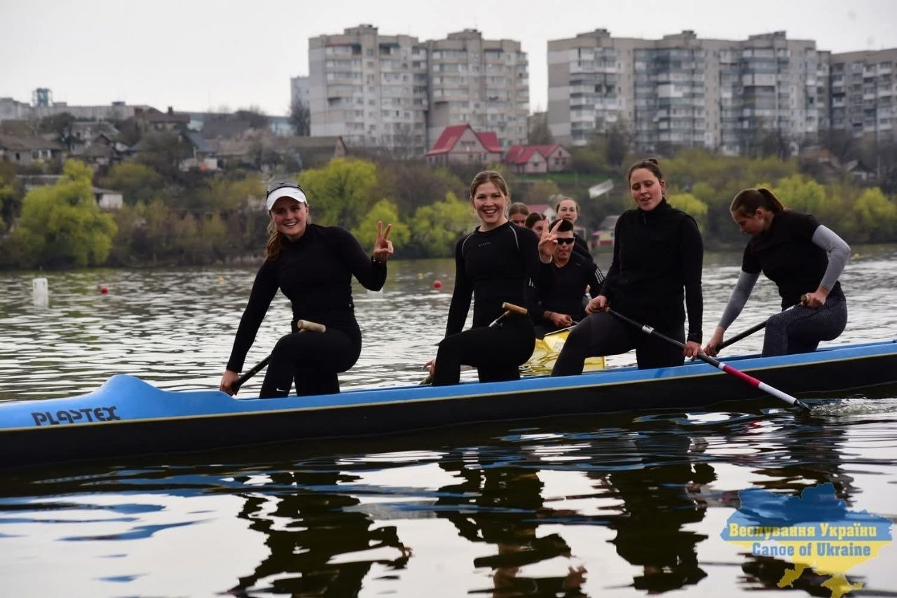 Група веслувальниць у чорному спортивному одязі позує у багатомісному каное на річці. Дівчата посміхаються, тримаючи весла, на фоні міської забудови та дерев.