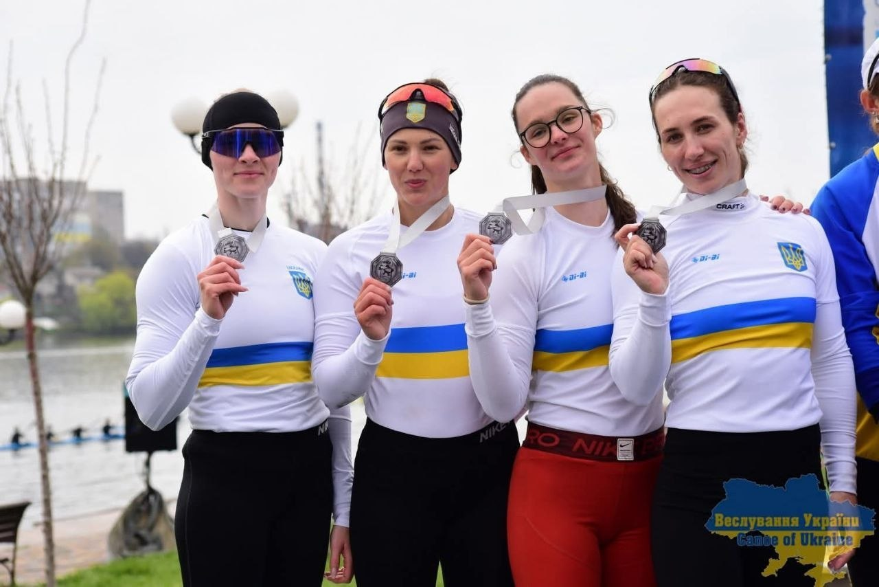 Чотири спортсменки у білій формі з українською символікою стоять поруч і демонструють здобуті срібні медалі. Вони посміхаються на фоні водойми, де тривають змагання з веслування.