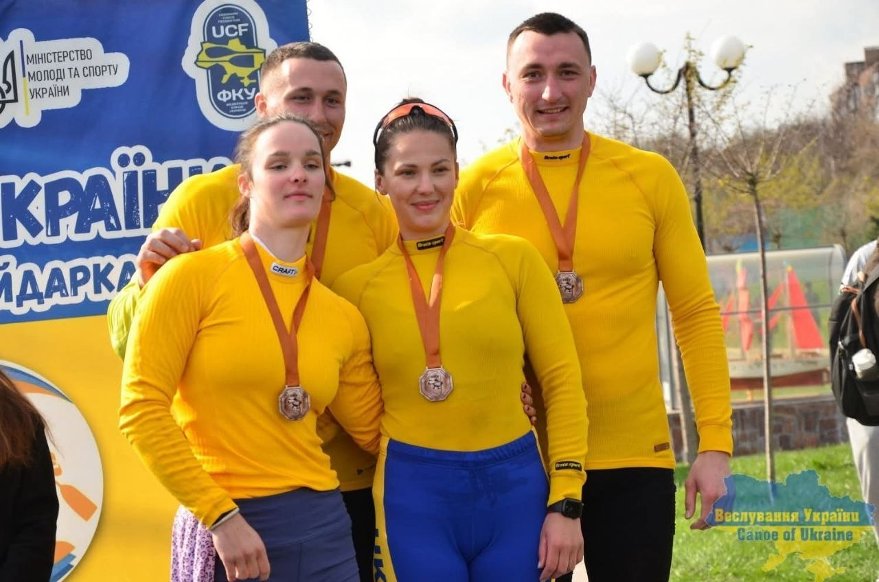 Двоє юнаків та двоє дівчат у жовтій спортивній формі позують із медалями на шиї на фоні банера змагань та паркової зони.