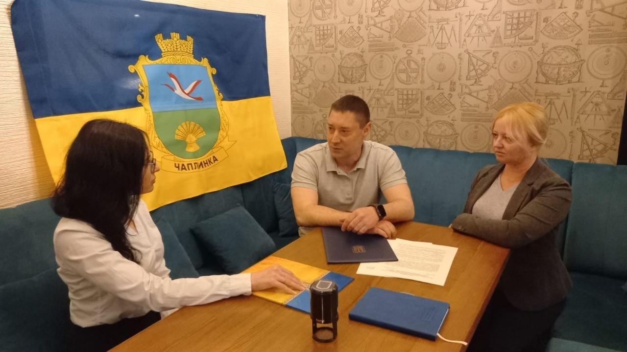 На зображенні троє людей — двоє жінок і чоловік — сидять за столом у приміщенні та обговорюють документи. На стіні за ними висить синьо-жовтий прапор із гербом селища Чаплинка. На столі лежать папки, папери та штамп.