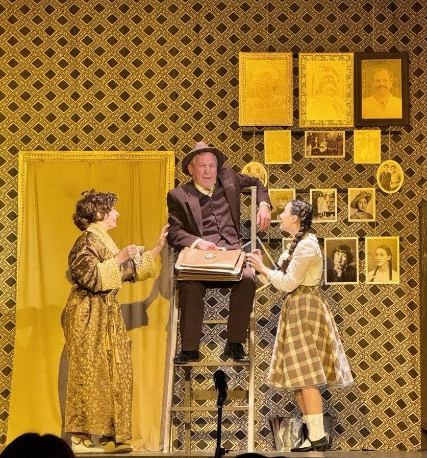 Театральна сцена: чоловік із портфелем сидить на драбині, а з обох боків до нього звертаються дві жінки в сценічних костюмах. Тло оздоблене візерунчастими шпалерами та багатьма портретами в рамках.
