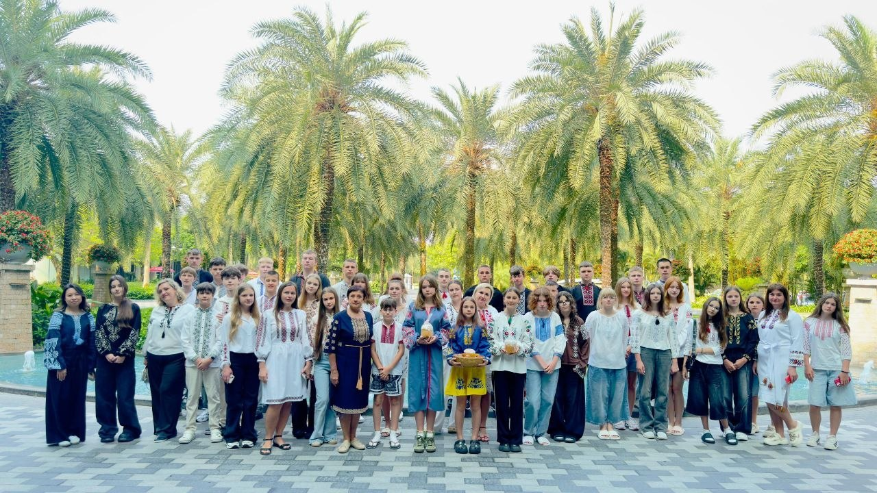 Група молодих людей та дорослих у вишиванках стоїть на відкритому майданчику, вимощеному плиткою. На задньому плані височіють численні пальми, а з боків видно фонтани. У центрі групи троє людей тримають традиційні українські калачі (короваї).