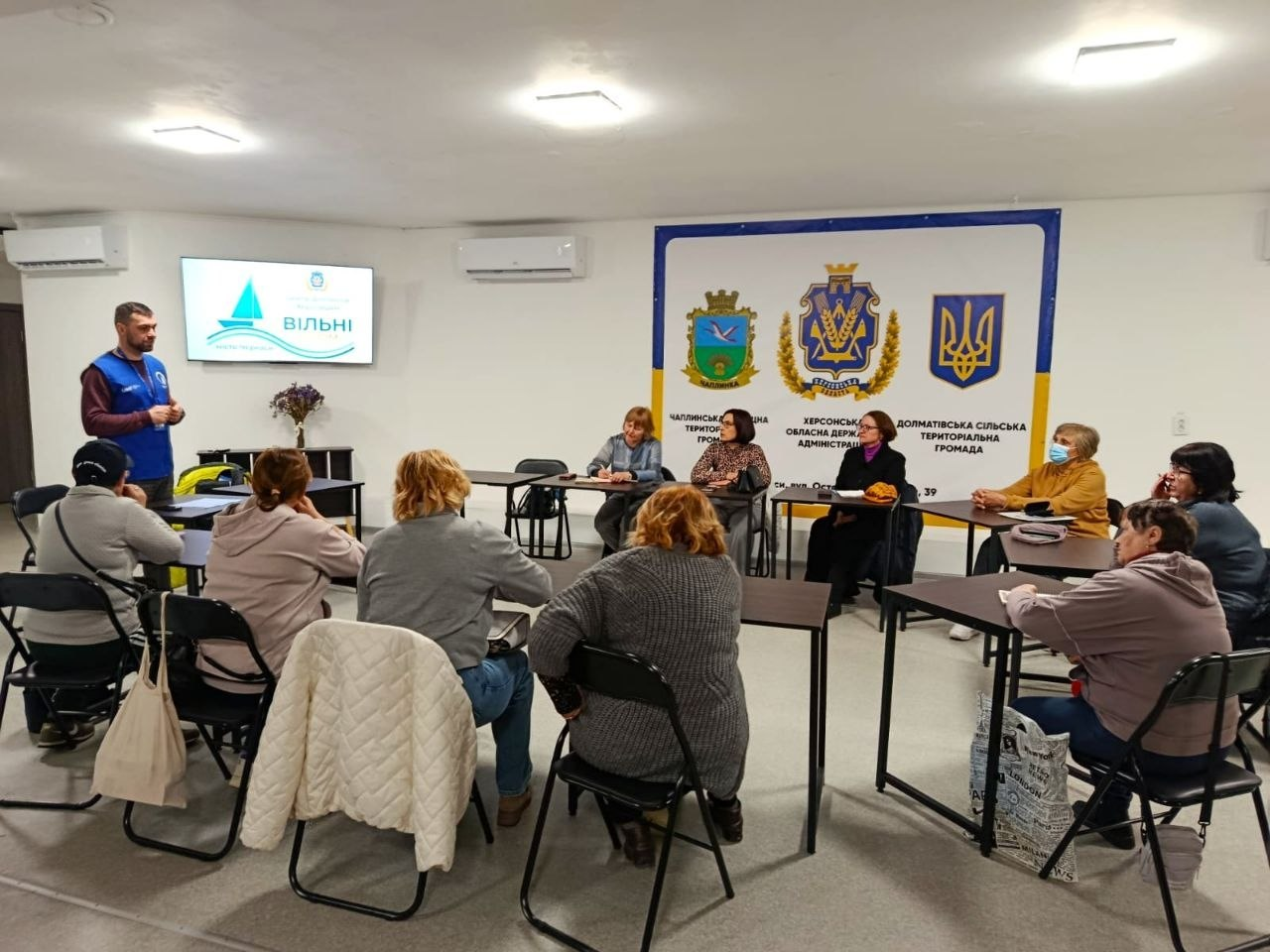 Робоча зустріч у приміщенні: група людей сидить за столами, розставленими півколом, і слухає чоловіка, який стоїть біля екрана з логотипом. На стіні розміщено великий банер із гербами та назвами територіальних громад і державних адміністрацій.