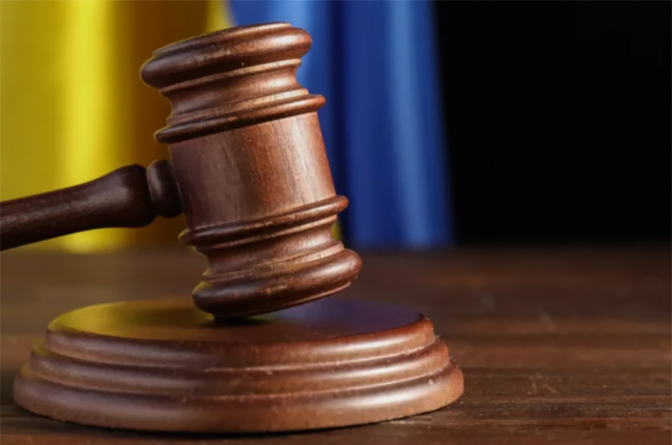 Дерев'яний суддівський молоток стоїть на підставці на столі. На розмитому фоні видно кольори українського прапора.