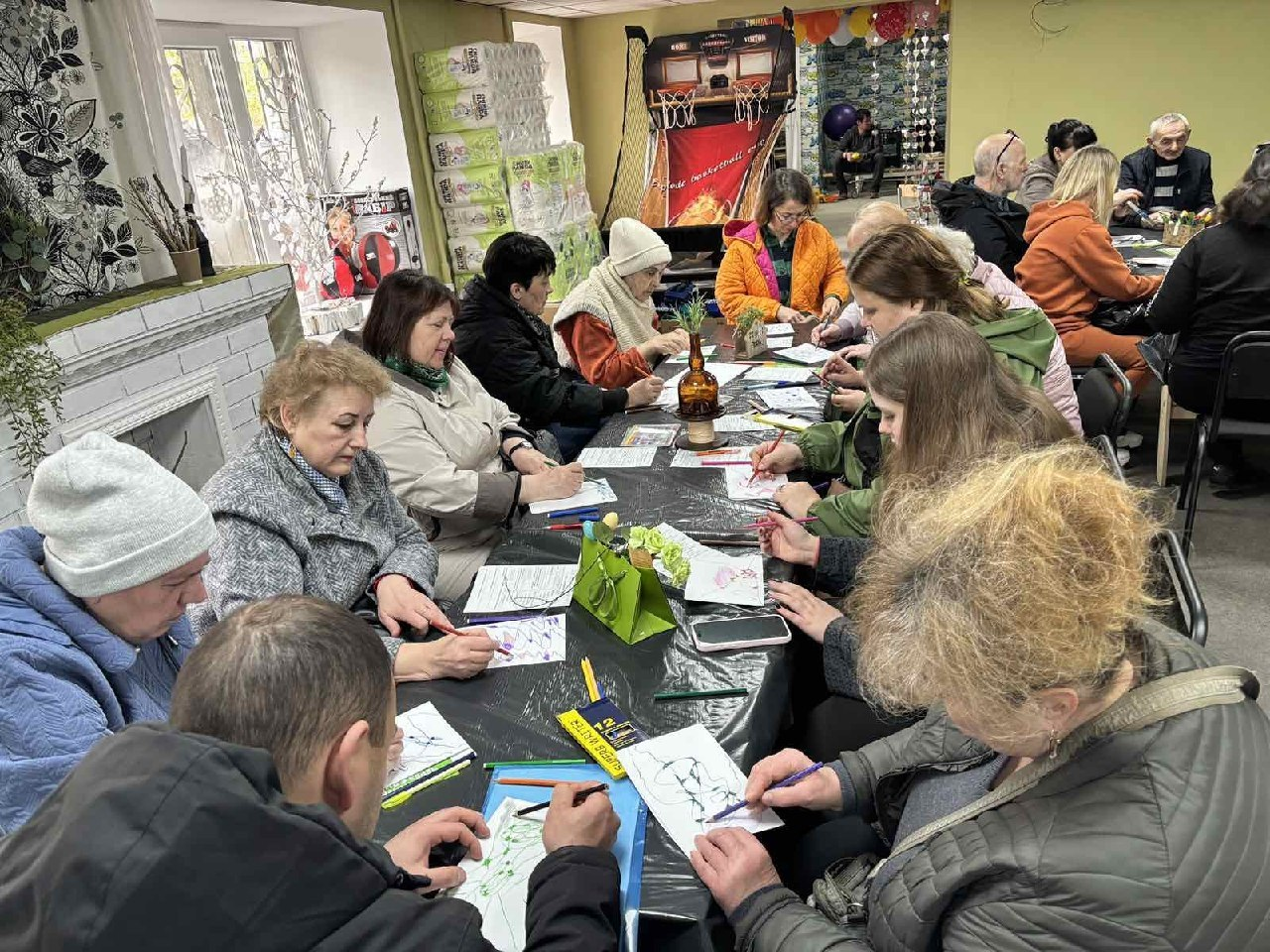 Група людей різного віку сидить за довгим столом у приміщенні та займається творчістю, малюючи олівцями на аркушах паперу. Навколо видно предмети побуту, іграшки та пакунки з гуманітарною допомогою.