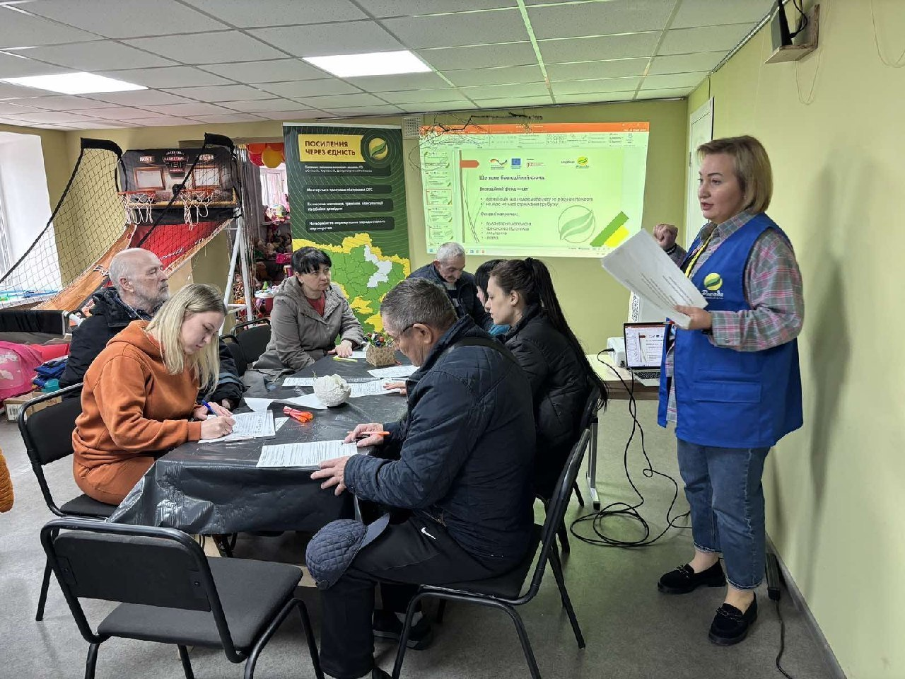 Жінка у синьому жилеті виступає перед групою людей, які сидять за столом у приміщенні. На фоні працює проєктор із презентацією та стоїть банер із заголовком «Посилення через єдність».