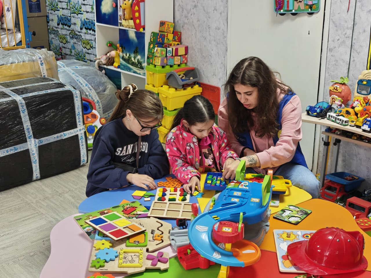 Жінка та двоє дівчаток сидять за столом у ігровій кімнаті, заповненій іграшками. Вони разом складають пазли та граються з розвивальними наборами.