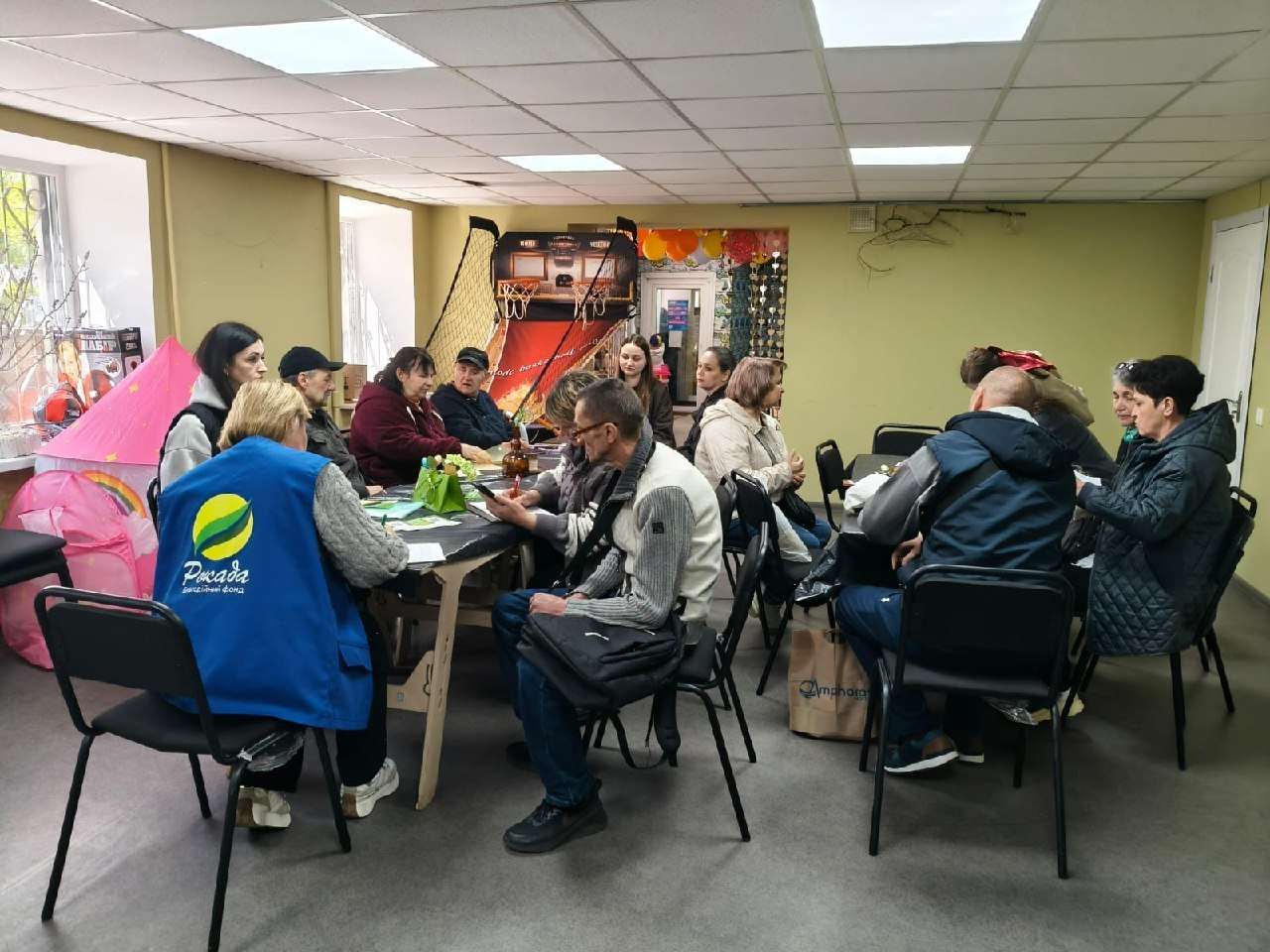 Група людей сидить за великим спільним столом у приміщенні, схожому на громадський чи волонтерський центр. Ліворуч на спинці стільця висить синій жилет із логотипом, а на фоні видно ігровий автомат для баскетболу та дитячий намет.