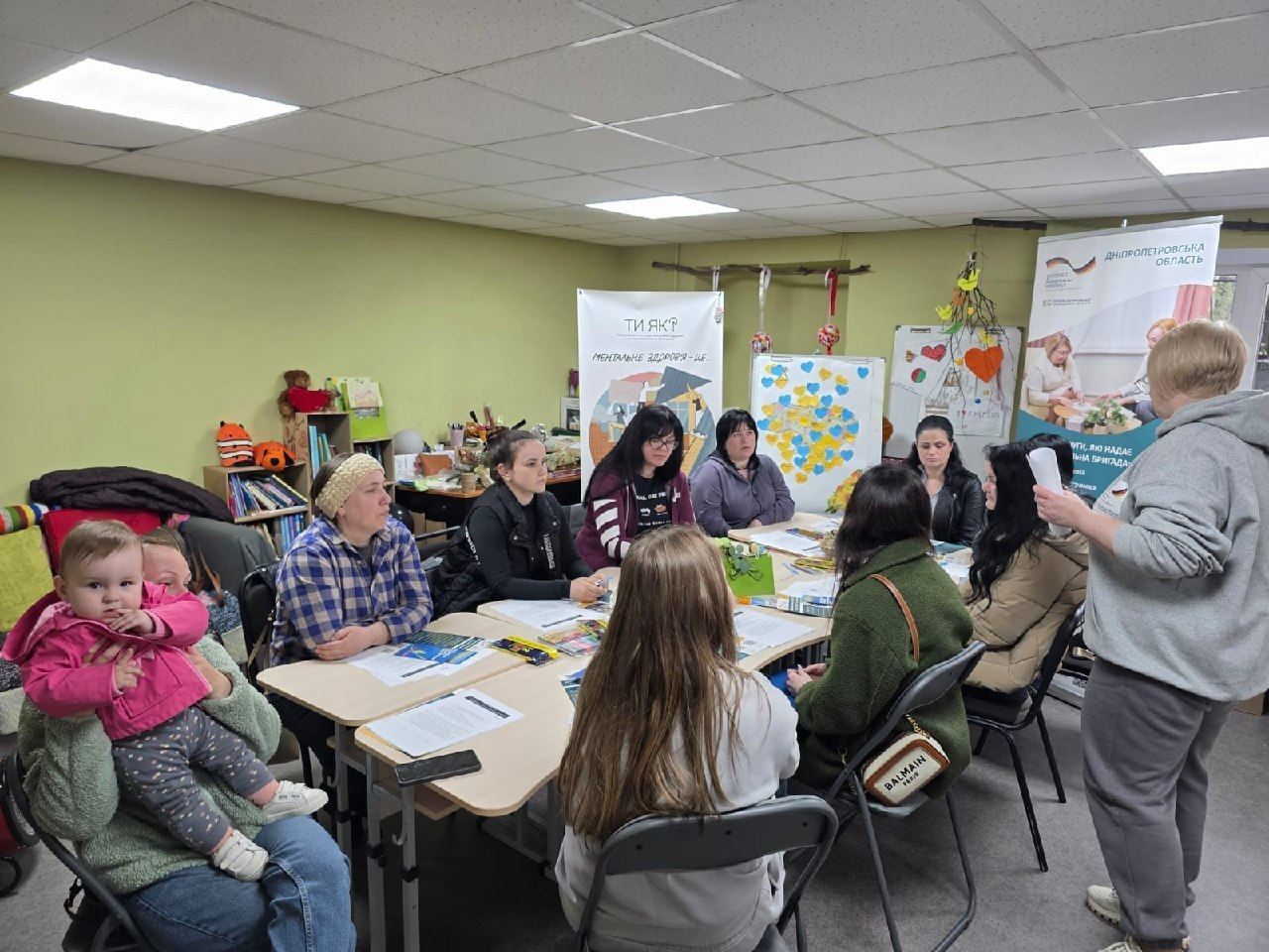 Група жінок із маленькою дитиною зібралася за столом у приміщенні для обговорення. На фоні стоять інформаційні банери, зокрема один із написом «Ти як? Ментальне здоров'я», а також дошка з паперовими сердечками кольорів українського прапора.
