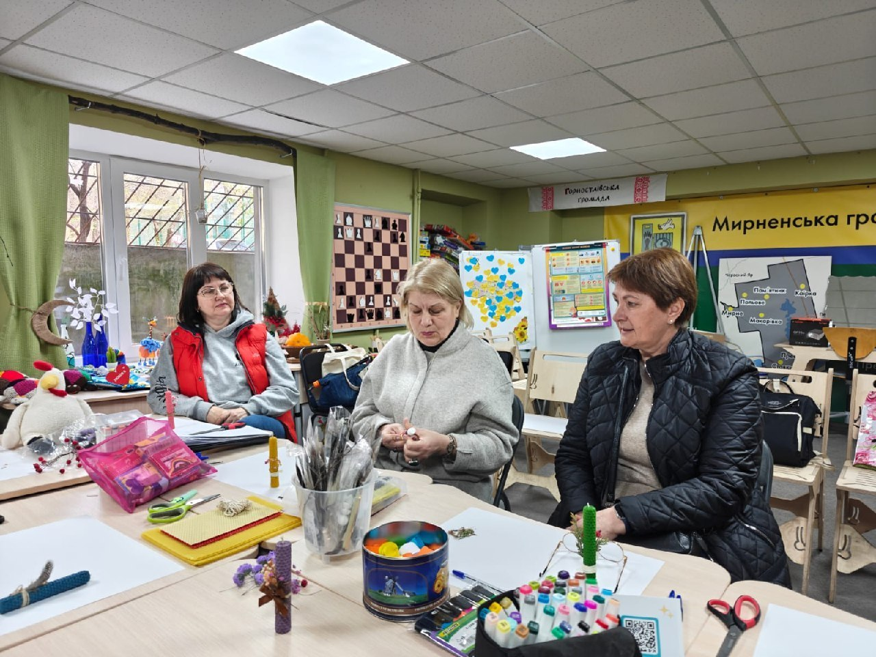 Три жінки сидять за столом у приміщенні, де на стінах видно карту Мирненської громади, шахову дошку та патріотичний стенд із сердечками. На столі розкладено матеріали для рукоділля: свічки, вощину, маркери та ножиці.