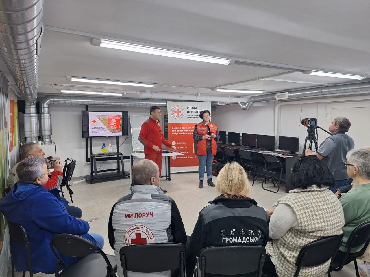 Тренінг з мінної безпеки: інструктори Червоного Хреста проводять навчання для групи людей у спеціально облаштованому приміщенні. На фоні встановлено інформаційний банер та екран із попереджувальними знаками.