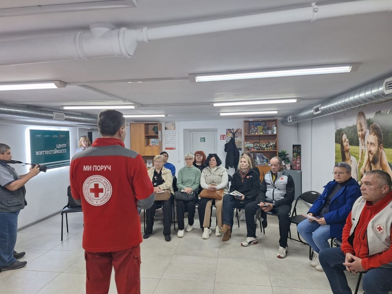 Представник Червоного Хреста у фірмовому жилеті стоїть спиною до камери та виступає перед групою людей, які сидять у «Центрі життєстійкості». Приміщення світле, обладнане під волонтерський хаб, а на стіні розміщено велике фото усміхненої сім'ї.