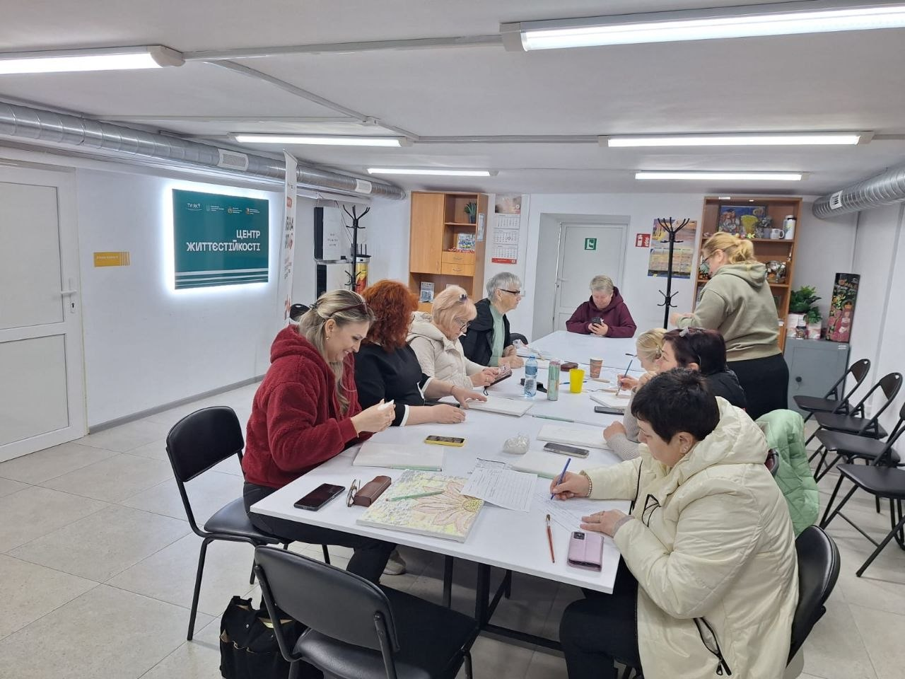 Група жінок сидить за довгим столом у світлому приміщенні «Центру життєстійкості». Вони зосереджено працюють з документами або займаються творчістю, а на стіні позаду них розміщено логотип центру з підсвіткою.