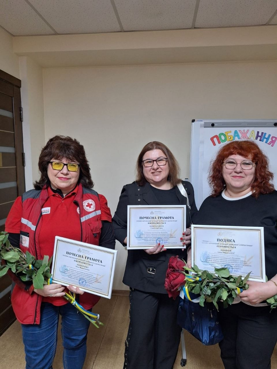 Три жінки стоять поруч у приміщенні та тримають у руках нагороди — «Почесні грамоти» та «Подяку», а також букети троянд, перев’язані синьо-жовтими стрічками. Жінка ліворуч одягнена у жилет із емблемою Червоного Хреста.