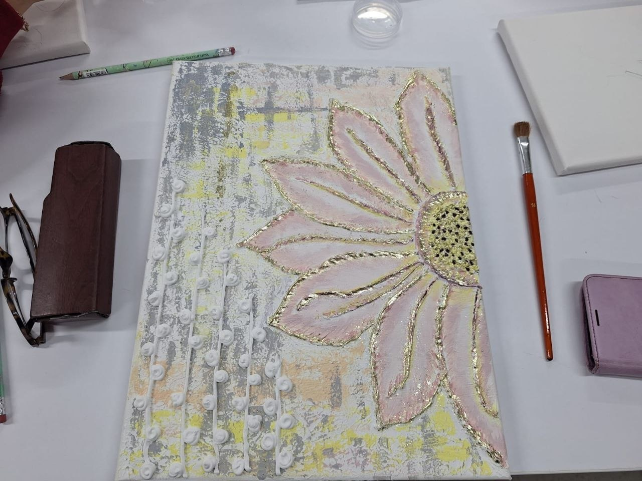 Картина на полотні, що лежить на білому столі, зображує велику стилізовану квітку з рожевими пелюстками та золотистим контуром. Поруч знаходяться художні інструменти: пензель, олівець, а також футляр для окулярів і мобільний телефон.