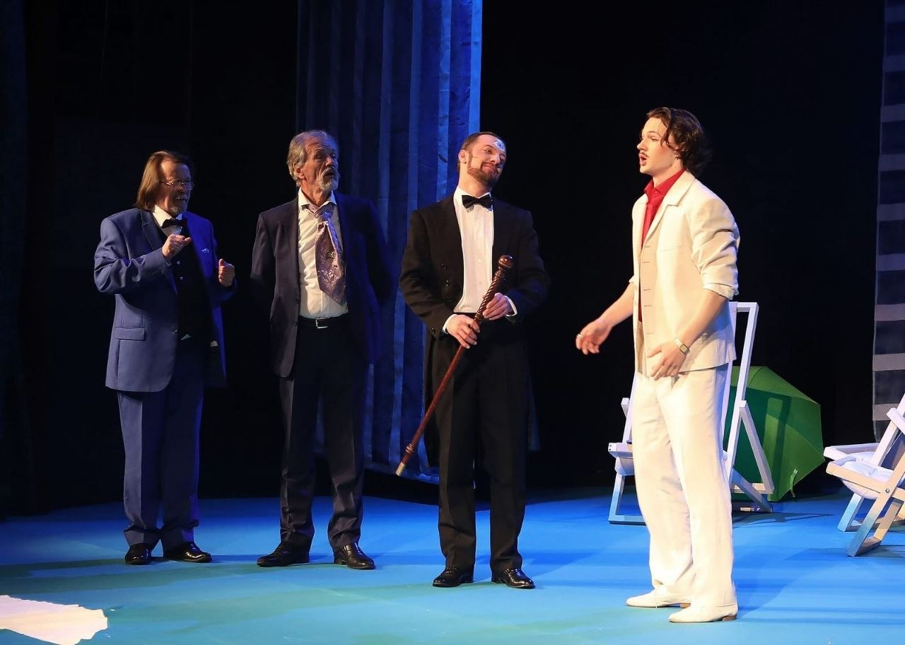 Чотири чоловіки в офіційних костюмах на театральній сцені під час вистави. Кожен має свій образ: від класичного смокінга з тростиною до яскравого білого костюма. Вони застигли в живій взаємодії на тлі темних декорацій.