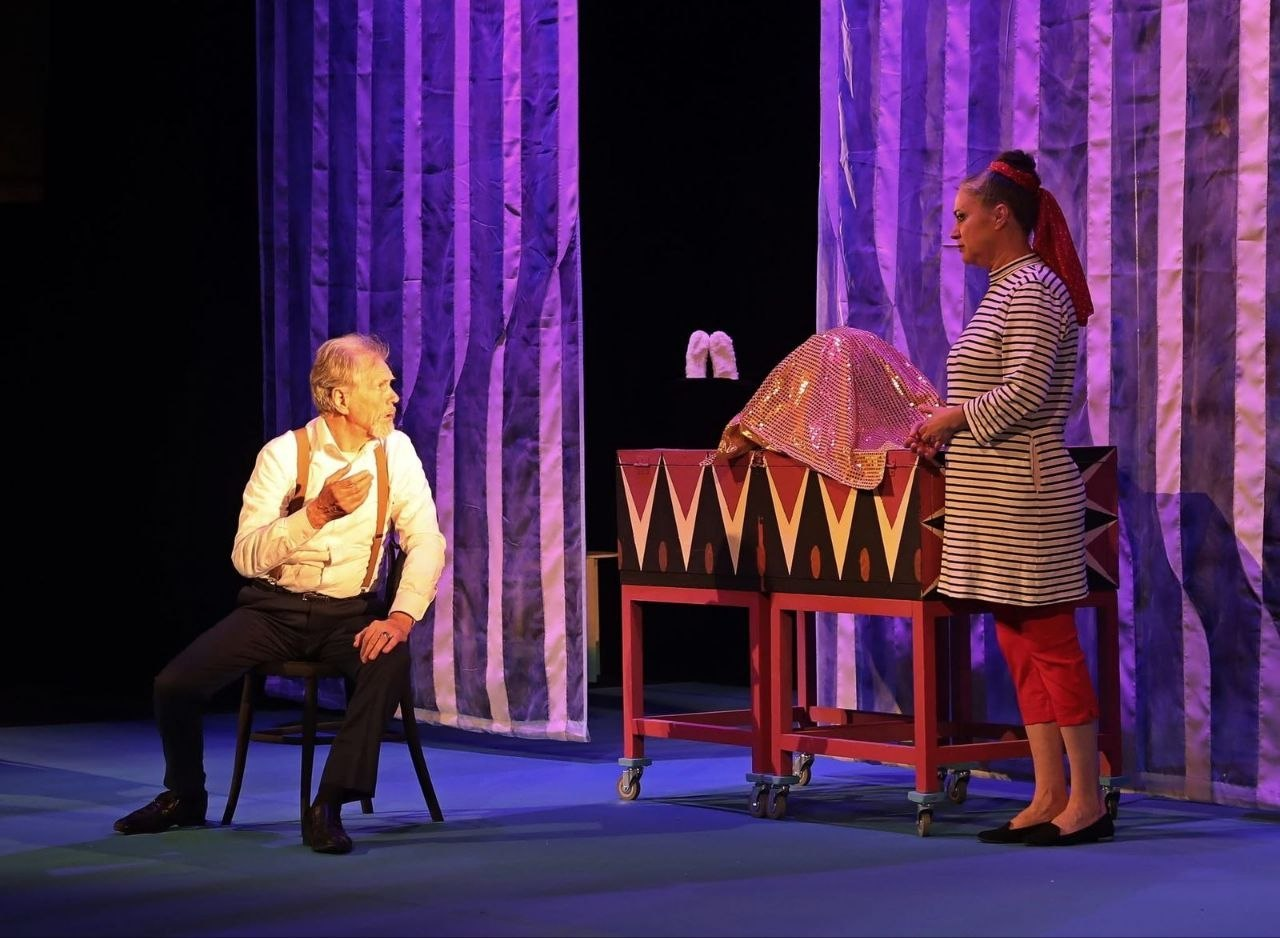 Сцена з театральної вистави: ліворуч на стільці сидить чоловік у світлій сорочці з підтяжками, емоційно притиснувши руку до грудей. Праворуч стоїть жінка у смугастій сукні та червоній хустці біля реквізиту, що нагадує ящик для фокусів. На задньому плані — синьо-білі смугасті завіси.