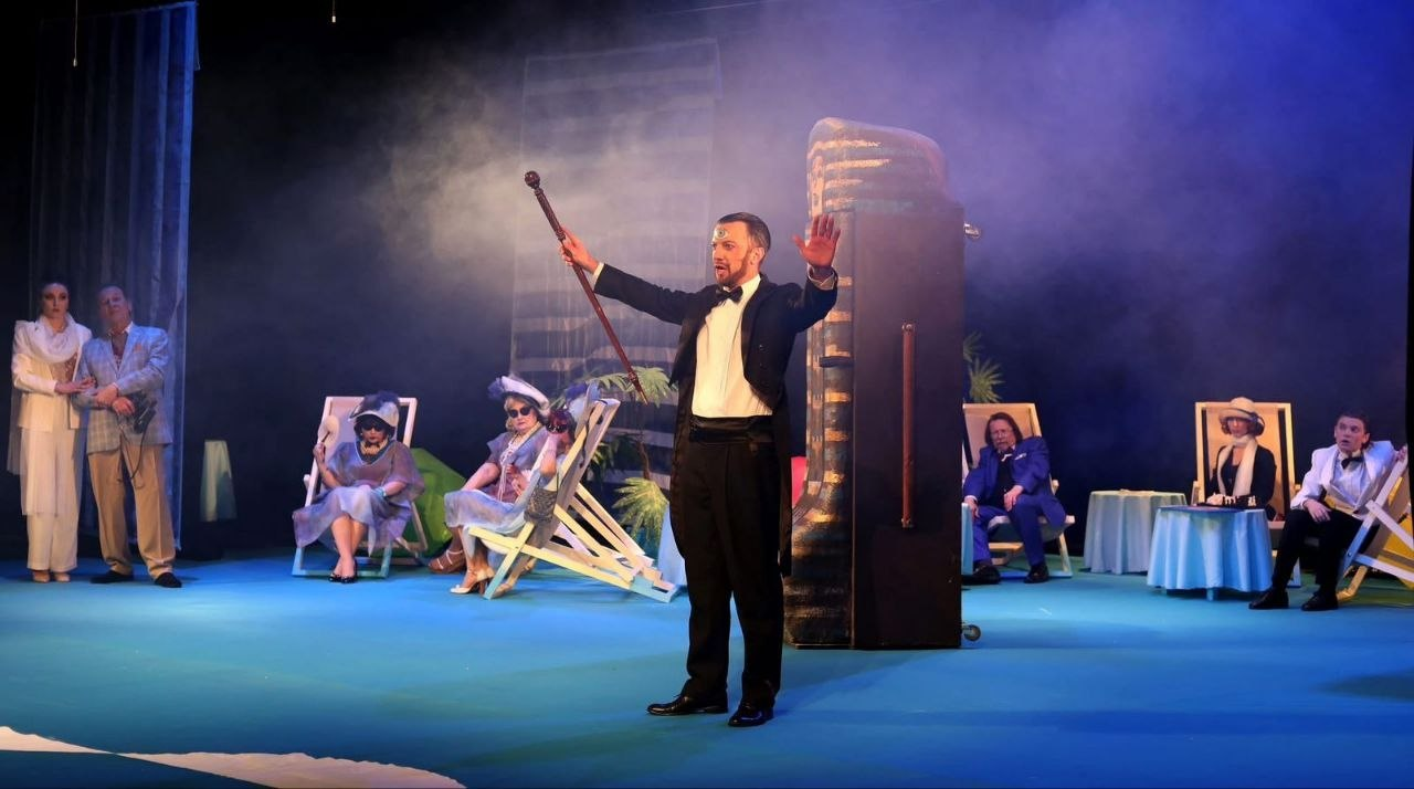 Театральна мізансцена: у центрі чоловік у смокінгу з піднятою тростиною, навколо якого на шезлонгах та біля столів розташувалися інші актори. Дія відбувається на фоні димової завіси та декорацій, що нагадують морський відпочинок.