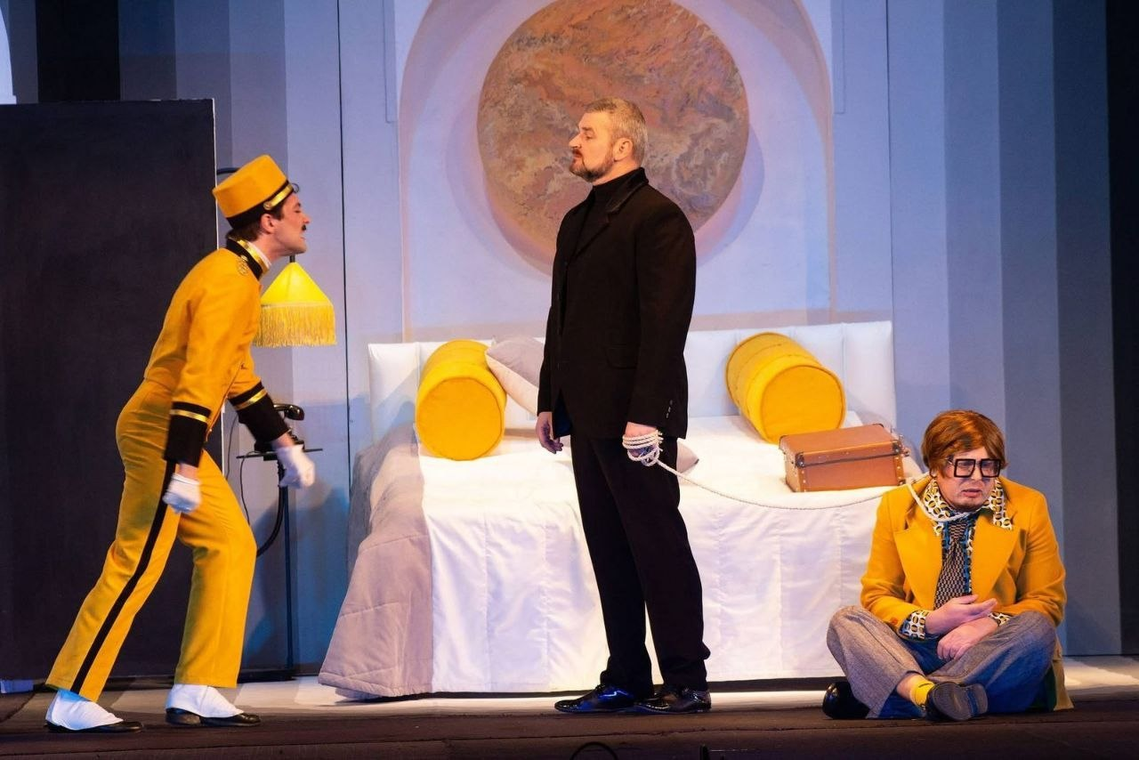 Сцена з вистави в готельному номері: зліва стоїть експресивний коридорний у яскраво-жовтій уніформі. У центрі — чоловік у чорному костюмі, який тримає на мотузці іншого чоловіка в жовтому піджаку, що сидить на підлозі. На задньому плані — ліжко з жовтими подушками-валиками та кругла декорація на стіні.