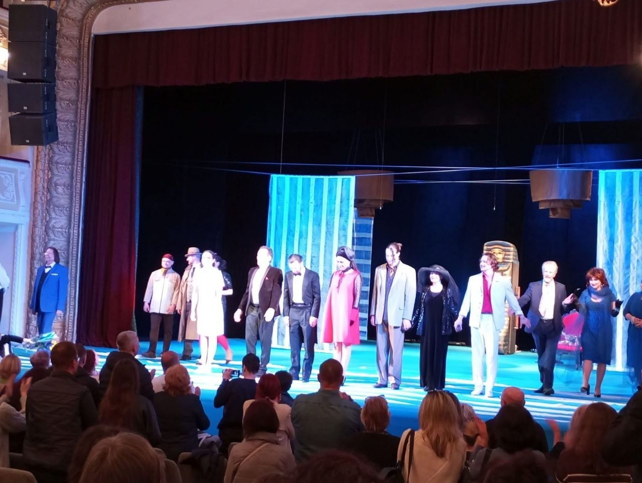 Театральна трупа в різноманітних костюмах виходить на уклін до глядачів після вистави. Актори стоять у ряд на яскраво освітленій сцені, а перед ними в залі видно глядачів, які аплодують. На задньому плані — сині декорації та масивна театральна завіса.