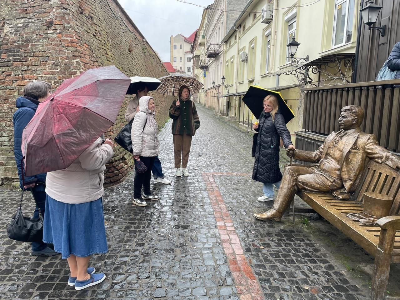 Екскурсійна група на вузькій брукованій вулиці біля пам'ятника Артуру Бернсу. Люди під парасольками слухають гіда на фоні старовинного муру та міської забудови.