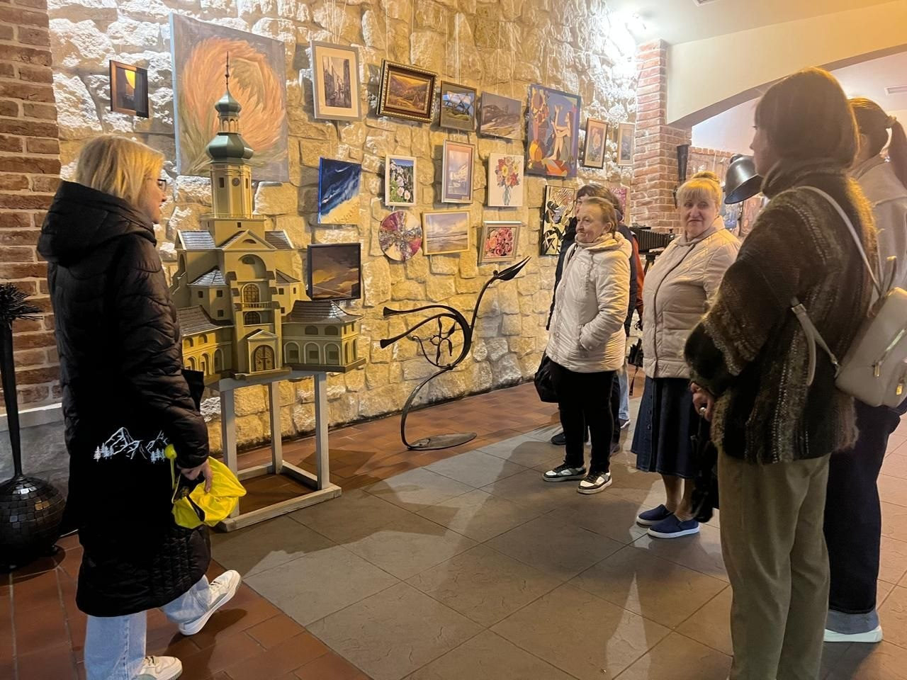 Група людей у мистецькому просторі розглядає виставку. У центрі уваги — деталізований макет старовинної будівлі з вежею, а на кам'яній стіні позаду розміщено багато різнопланових картин.