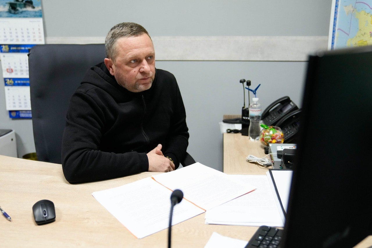 Чоловік у чорному худі сидить за робочим столом в офісі, зосереджено дивлячись на екран монітора. Перед ним лежать папери, на столі також видно мишку, мікрофон, настільний телефон та пляшку води. На стіні позаду висять календарі та мапа.