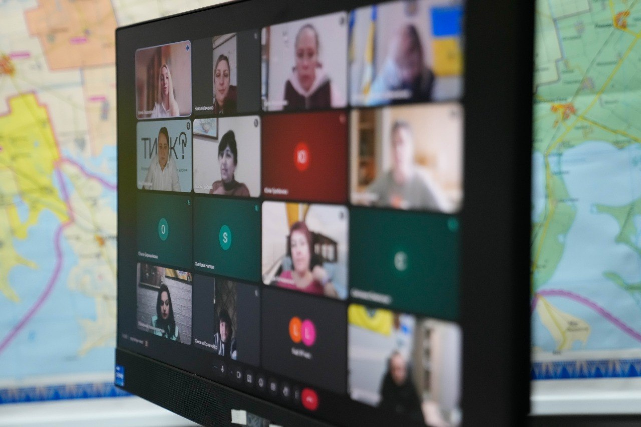 Чорний монітор комп'ютера, на екрані якого відкрито вікно відеоконференції з сіткою учасників. Один із учасників використовує віртуальний фон із логотипом програми ментального здоров’я «Ти як?». Монітор стоїть на фоні великої настінної карти, що додає кадру робочої та офіційної атмосфери.